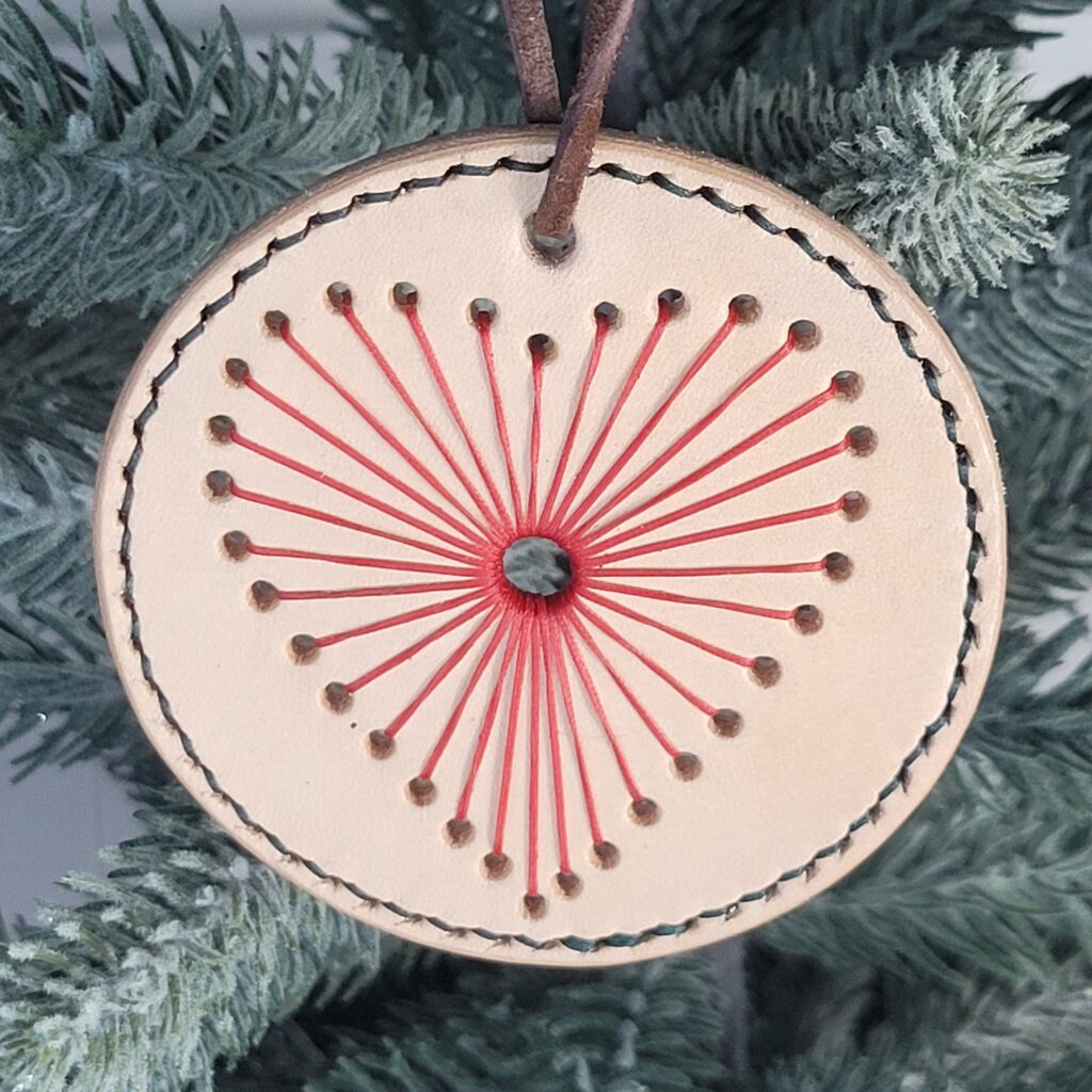 String heart ornament. A tan circle with a heart stitched in red. The heart has a hole in the middle with thread radiating out from the hole to make the heart shape. A line of green thread is stitched around the edge to make a border.