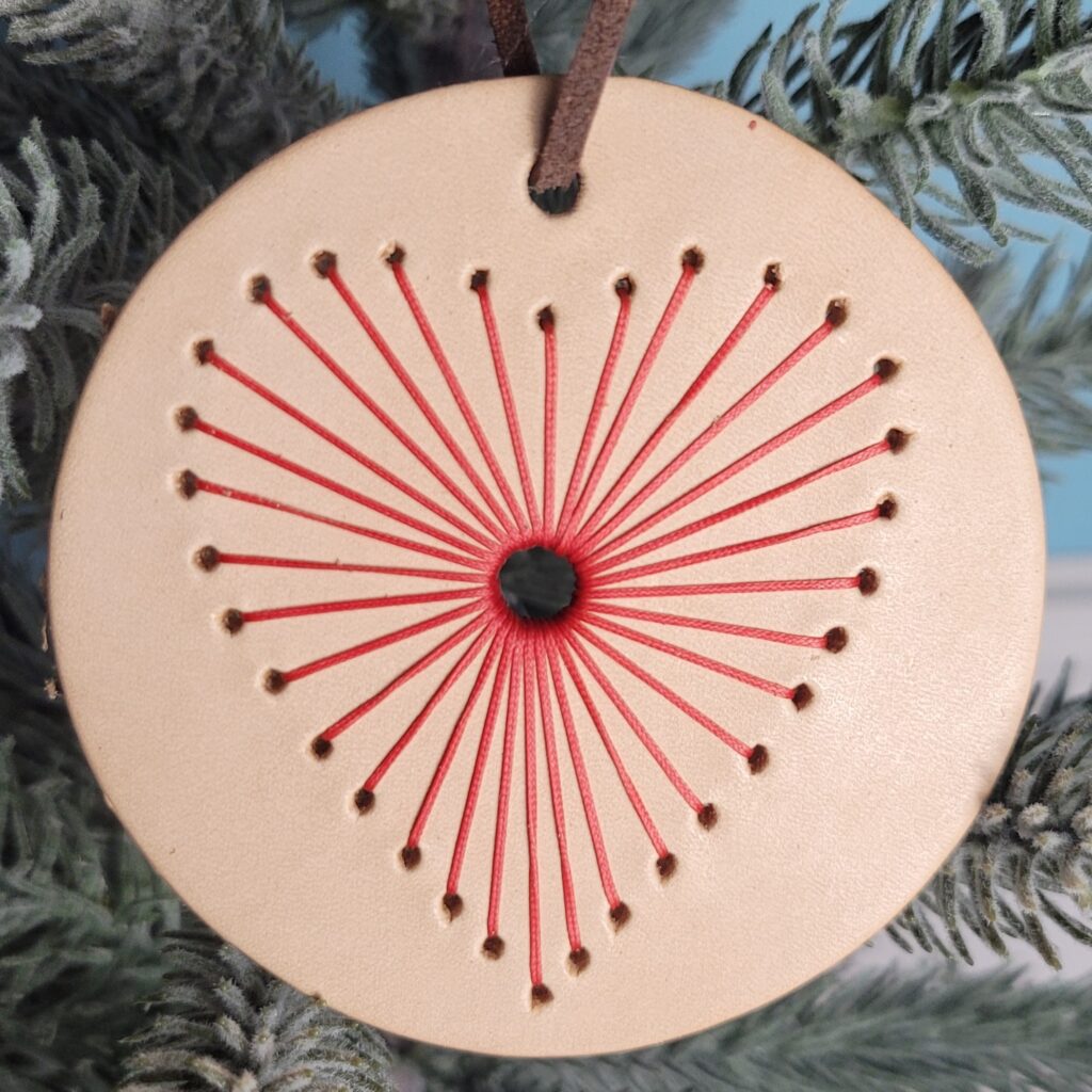 String heart ornament. A tan circle with a heart stitched in red. The heart has a hole in the middle with thread radiating out from the hole to make the heart shape.