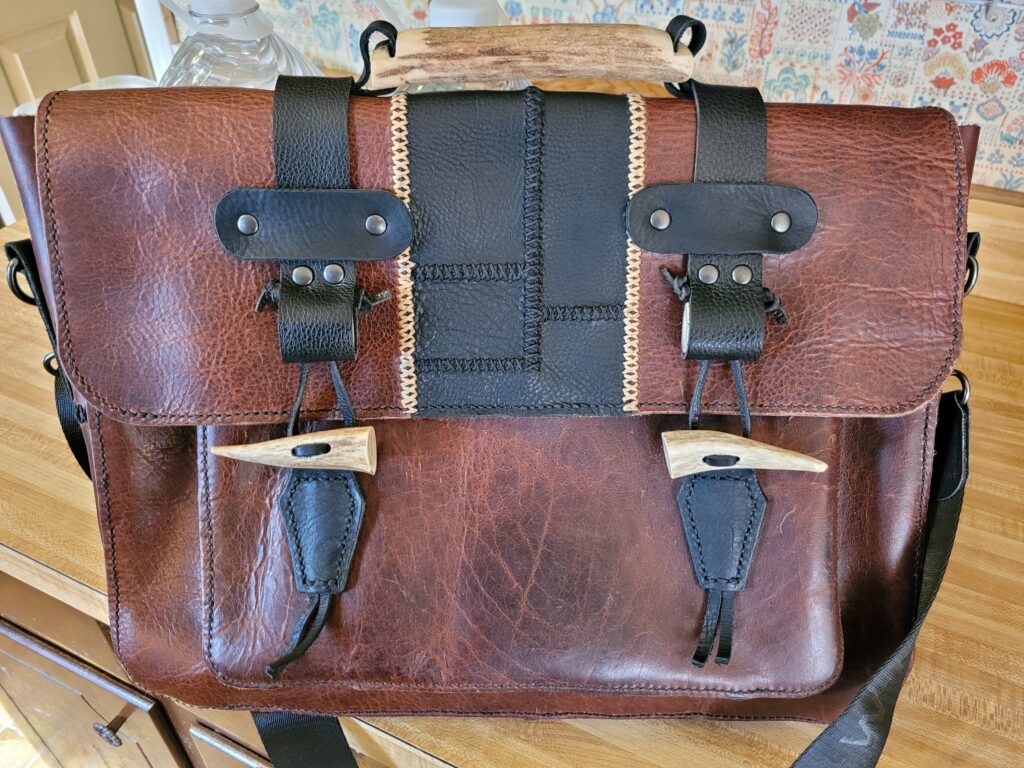 Brown messenger bag with patched black section on front flap. Black straps come over the top for closure. Closure toggles and top handle made of antler pieces