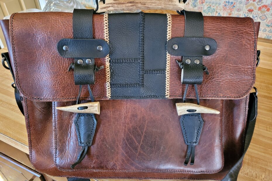 Brown messenger bag with patched black section on front flap. Black straps come over the top for closure. Closure toggles and top handle made of antler pieces